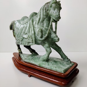 May include: A green jade horse sculpture on a wooden base. The horse is depicted in a galloping pose with a saddle and bridle.