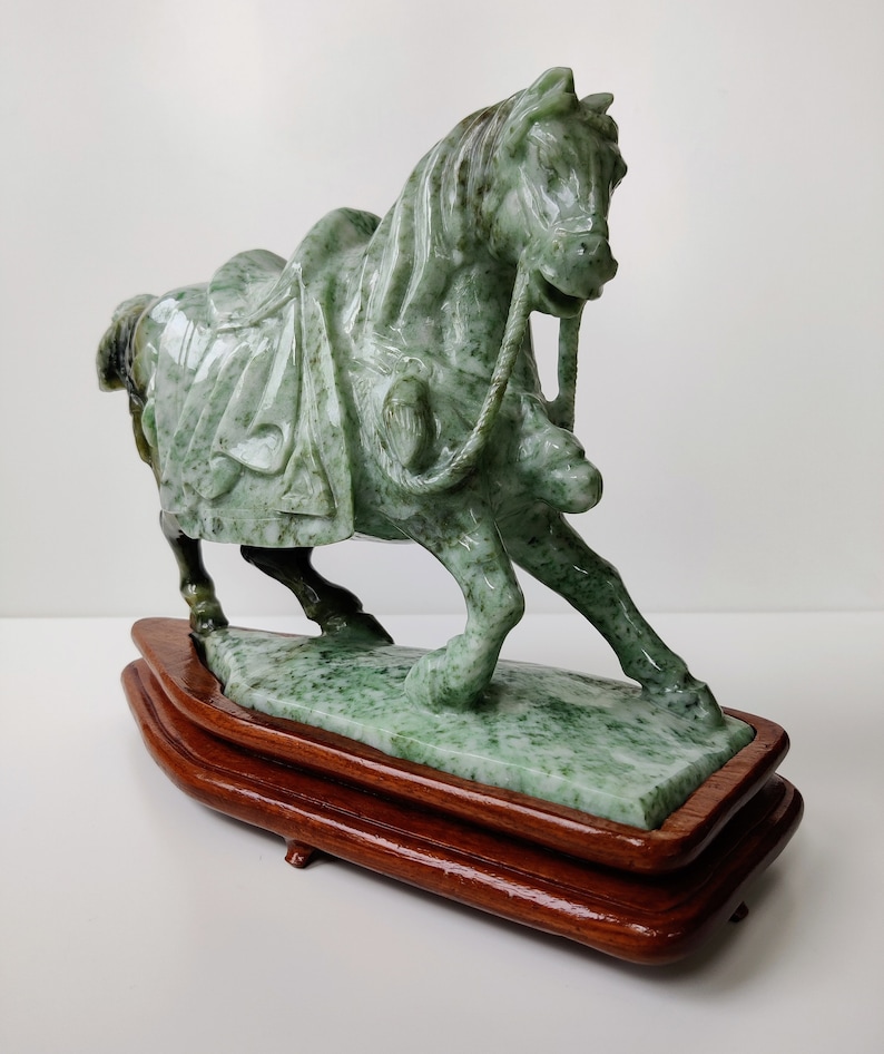 May include: A green jade horse sculpture on a wooden base. The horse is depicted in a galloping pose with a saddle and bridle.