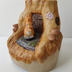 May include: A ceramic musical figurine of a squirrel standing on a tree stump with a small door and mushrooms. The squirrel is brown and has a pink flower on the stump.