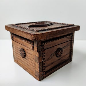 May include: A small, intricately carved wooden box with a hinged lid. The box is made of dark wood and has a decorative pattern around the edges. The lid is closed and has a carved design in the centre.