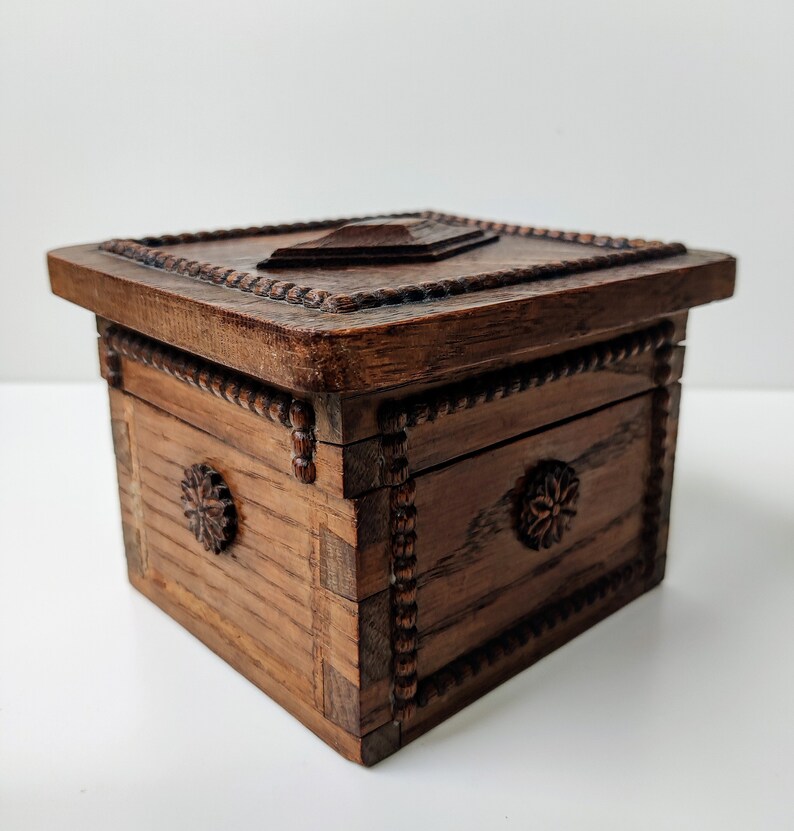 May include: A small, intricately carved wooden box with a hinged lid. The box is made of dark wood and has a decorative pattern around the edges. The lid is closed and has a carved design in the centre.