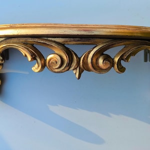 May include: A gold-painted wooden shelf with ornate scrollwork details. The shelf is curved and has a decorative design on the front.