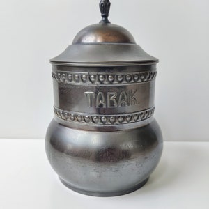 Puede incluir: Un tarro de tabaco de metal negro con una tapa adornada y la palabra "TABAK" en relieve en la parte delantera.