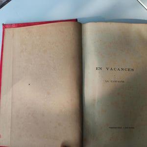 May include: Open vintage book with red binding. The pages are aged and show signs of wear. The text "EN VACANCES" and "LA CAMPAGNE" is visible on the right page. The book is open on a light blue surface.