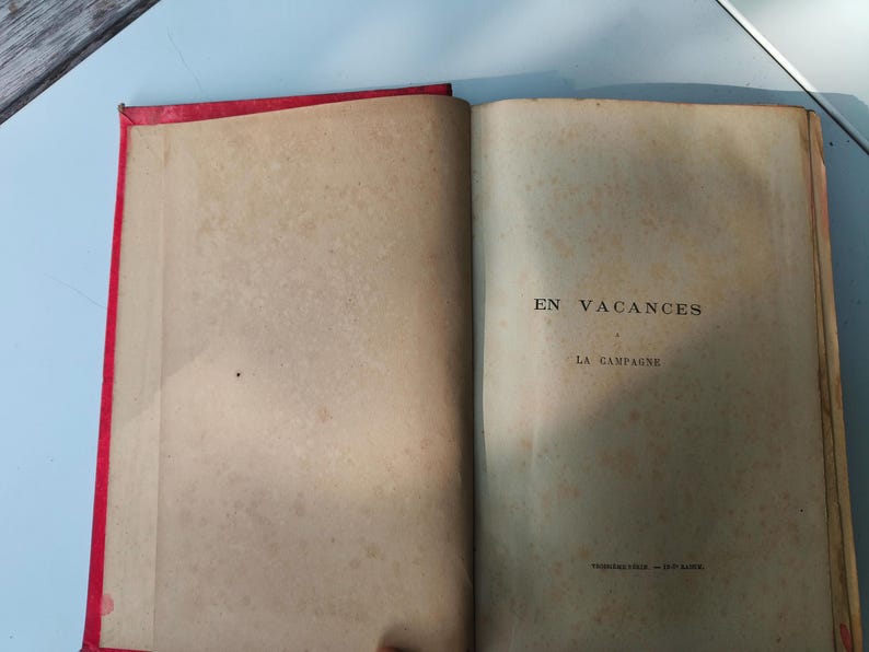 May include: Open vintage book with red binding. The pages are aged and show signs of wear. The text "EN VACANCES" and "LA CAMPAGNE" is visible on the right page. The book is open on a light blue surface.