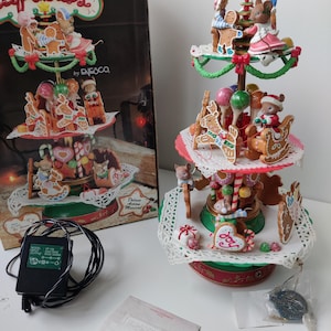 May include: A three-tiered gingerbread carousel with a Christmas theme. The carousel features gingerbread houses, candy canes, and a gingerbread man. The carousel is lit up with lights and plays music. The box says "Sugar N Spice Capades To Be Home For Christmas".