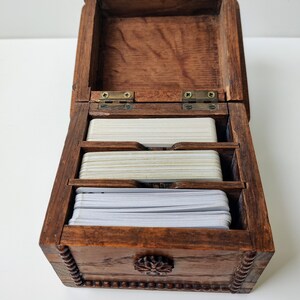 May include: A wooden box with a hinged lid and a carved floral design on the front. The box is open and contains three stacks of white cards.