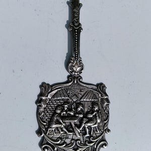 Pode incluir: Um servidor de bolo prateado com um design ornamentado. O cabo apresenta um grupo de pessoas em relevo, enquanto a lâmina representa uma cena de pessoas reunidas em torno de uma mesa.