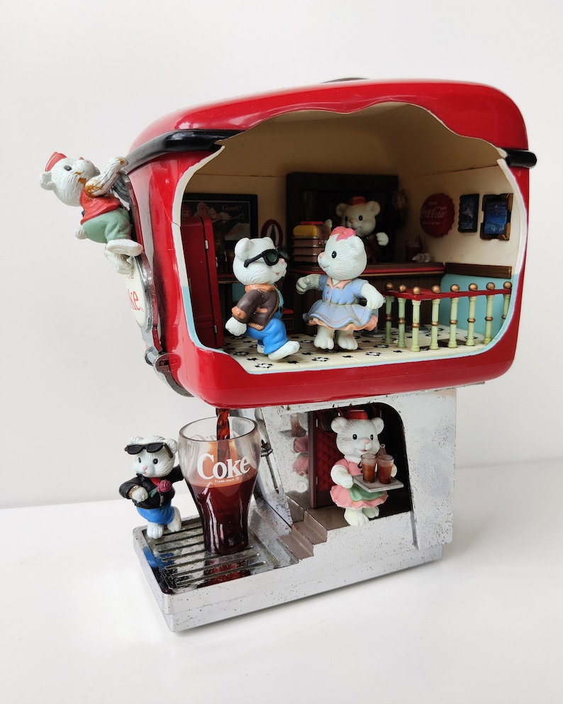 May include: A red Coca-Cola themed miniature diner with a white roof and a silver base. The diner has a window with a Coca-Cola sign and a small interior with five white polar bear figurines. One polar bear is climbing a ladder, one is serving drinks, and three are inside the diner. The diner is pouring a glass of Coca-Cola.