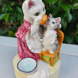 May include: A ceramic ornament featuring two cat figures. One cat, dressed in pink and white, offers a small, round item to a smaller cat on a miniature chair. A small bowl with a blue interior is also visible.