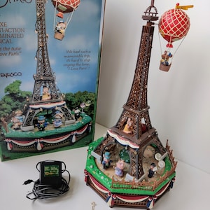 May include: A musical carousel featuring a miniature Eiffel Tower with a hot air balloon and figurines of mice. The carousel is decorated with red, white, and blue bunting. The box includes the text "I Love Paris".