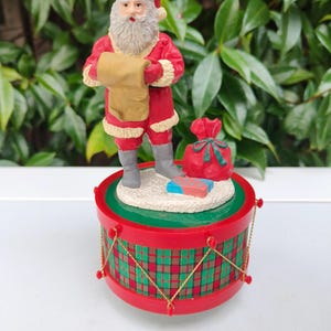 Puede incluir: Figura navideña de Papá Noel con traje y gorro rojos, sosteniendo un pergamino. Se encuentra sobre una base con forma de tambor con un patrón de cuadros rojos y verdes. También hay un saco rojo con un lazo verde.
