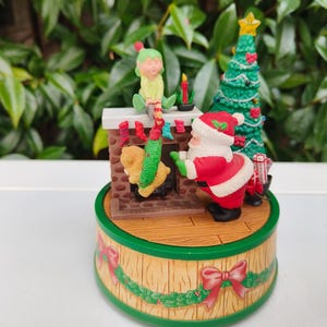 Puede incluir: Figura navideña con Papá Noel, un elfo y un árbol de Navidad decorado. Papá Noel, con traje rojo, se acerca a una chimenea. La base está decorada con guirnaldas verdes y lazos rojos. La figura mide unos 15 cm de alto.