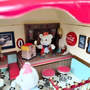 May include: A miniature diner scene with a teddy bear wearing a red and white uniform serving drinks at the counter. The diner has a red and white striped awning, a jukebox, and a Coca-Cola sign.