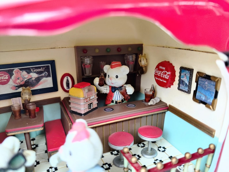 May include: A miniature diner scene with a teddy bear wearing a red and white uniform serving drinks at the counter. The diner has a red and white striped awning, a jukebox, and a Coca-Cola sign.