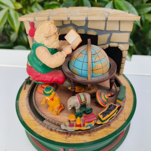 Puede incluir: Un carrusel musical temático navideño con Papá Noel leyendo un libro junto a un globo terráqueo. El carrusel incluye juguetes en miniatura como un tren, un elefante y una peonza. La base es verde con detalles rojos, creando un ambiente festivo.