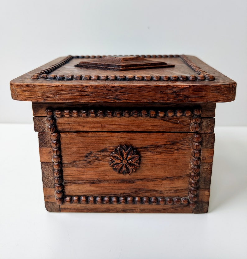 May include: A wooden box with a carved floral design on the front and a beaded border around the edges. The box has a hinged lid with a carved design on top.