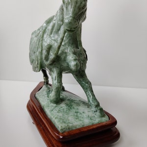 May include: A green jade horse sculpture on a wooden base. The horse is depicted in a running pose with its head turned to the side. The sculpture is detailed with intricate carvings.