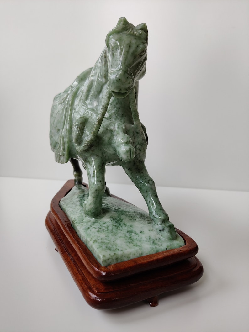 May include: A green jade horse sculpture on a wooden base. The horse is depicted in a running pose with its head turned to the side. The sculpture is detailed with intricate carvings.