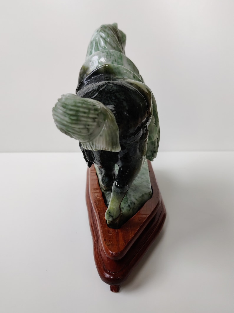 May include: A green jade horse sculpture on a wooden stand. The horse is carved in a standing pose with its head turned to the side. The jade is a mottled green colour with black and white streaks.