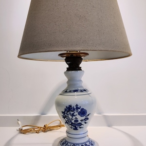 Puede incluir: Una lámpara de mesa de porcelana blanca con un diseño floral azul y una pantalla de lino beige.