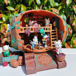 May include: A whimsical wooden wine barrel playset with a brown, green and white colour scheme. The barrel is decorated with vines and grapes. The playset features a small wooden door, a ladder and several small figurines of mice. The text "The Winery" is printed on the front of the barrel.