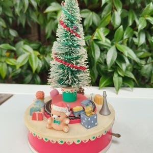 Puede incluir: Un pequeño árbol de Navidad verde esmerilado, adornado con una guirnalda de cuentas rojas y una estrella amarilla. La base es un carrusel musical con figuras de juguete, incluyendo un oso de peluche con gorro de Papá Noel y regalos en miniatura.