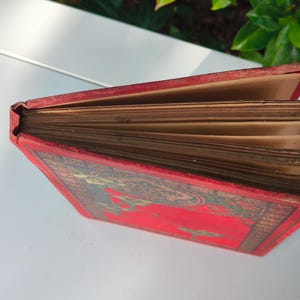 May include: An antique book with a red cover and aged, yellowed pages. The cover features an ornate design with gold and green accents. The book's spine is visible, showing the binding and the wear of time. The pages are fanned out, revealing their age.