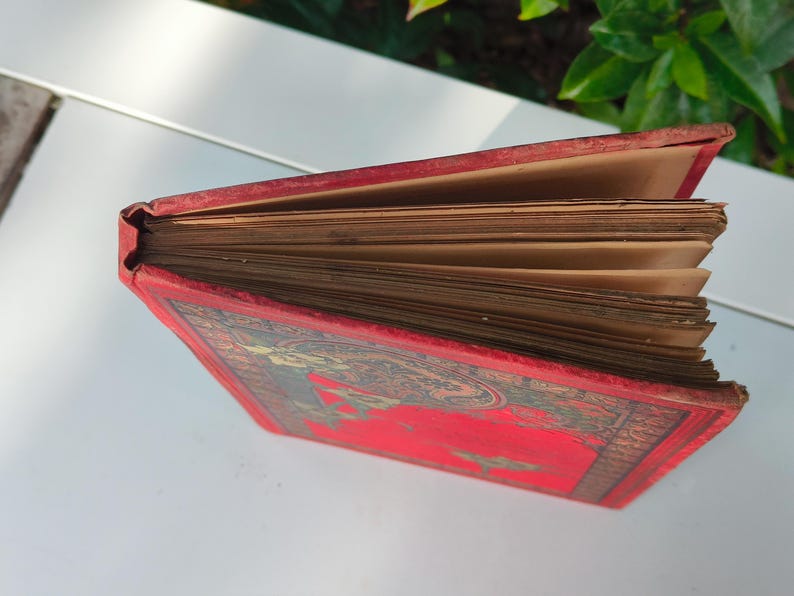May include: An antique book with a red cover and aged, yellowed pages. The cover features an ornate design with gold and green accents. The book's spine is visible, showing the binding and the wear of time. The pages are fanned out, revealing their age.