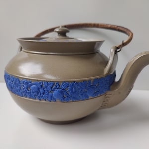 Puede incluir: Una tetera marrón con un diseño floral azul alrededor del medio. La tetera tiene un asa de mimbre marrón y una tapa.