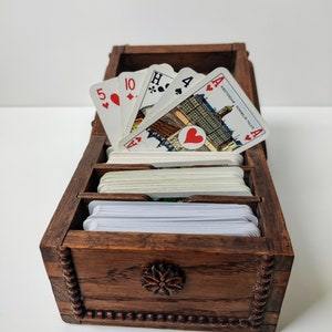 May include: A wooden box with a carved floral design on the front. The box is open and contains a pack of playing cards. The top cards are an Ace of Hearts, a 4 of Spades, a Jack of Hearts, and a 10 of Diamonds. The cards have a city scene on the back.