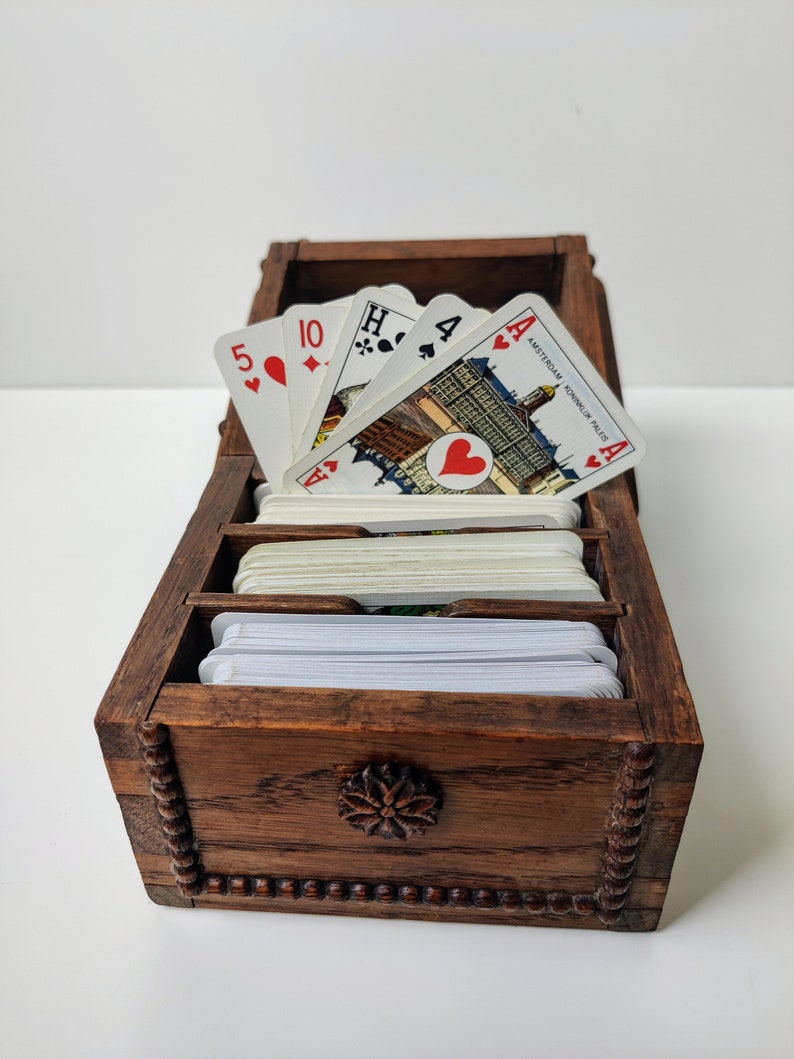 May include: A wooden box with a carved floral design on the front. The box is open and contains a pack of playing cards. The top cards are an Ace of Hearts, a 4 of Spades, a Jack of Hearts, and a 10 of Diamonds. The cards have a city scene on the back.