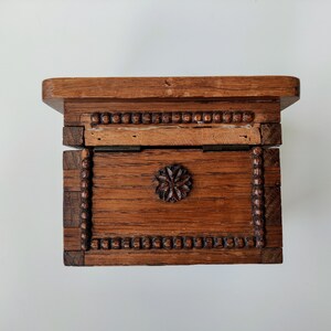 May include: A wooden box with a hinged lid and a carved floral design on the front. The box is made of dark wood and has a beaded trim around the edges.