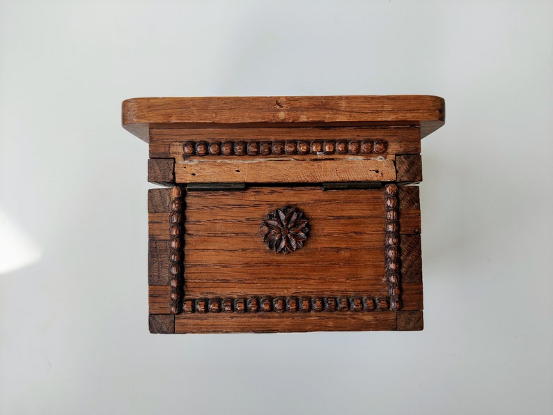 May include: A wooden box with a hinged lid and a carved floral design on the front. The box is made of dark wood and has a beaded trim around the edges.