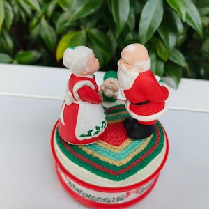 Puede incluir: Una figura navideña con Papá Noel, Mamá Noel y un elfo. Papá Noel y Mamá Noel se enfrentan, con el elfo entre ellos. La figura se encuentra sobre una base redonda con un diseño rojo, verde y blanco.