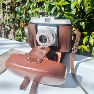 Puede incluir: Una cámara vintage plateada y negra con una funda y correa de cuero marrón. La lente de la cámara tiene el texto "ISOLA I" y marcas de medición. La funda está abierta, revelando la cámara, y la correa está sujeta.