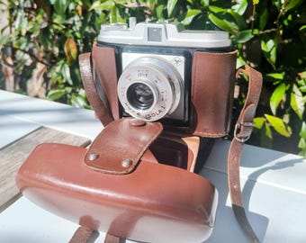 May include: A vintage silver and black camera with a brown leather case and strap. The camera lens has the text "ISOLA I" and measurement markings. The case is open, revealing the camera, and the strap is attached.