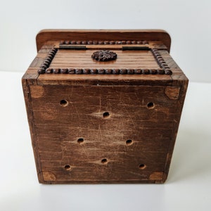 May include: A small wooden box with a carved design on the top. The box has a hinged lid and is made of dark brown wood. The box has a series of small holes on the bottom.