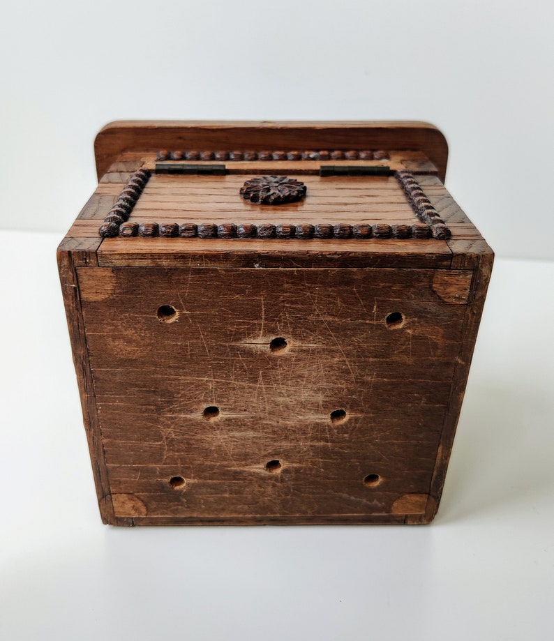 May include: A small wooden box with a carved design on the top. The box has a hinged lid and is made of dark brown wood. The box has a series of small holes on the bottom.