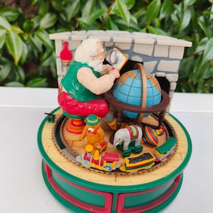 Puede incluir: Figura navideña que representa a Papá Noel examinando un globo terráqueo con una lupa. La escena incluye una chimenea, un tren de juguete y figuras de animales. La base es verde y roja, con un diseño circular.