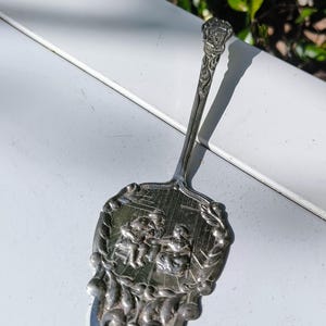 Pode incluir: Utensílio de servir prateado com um cabo decorativo e uma lâmina detalhada e ornamentada. A lâmina apresenta uma cena em relevo de figuras, emoldurada por uma borda floral. O cabo é fino e termina com um brasão detalhado.