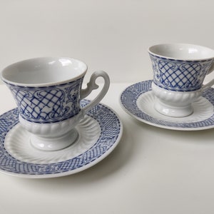 May include: Two white porcelain teacups with blue and white patterned designs. The cups have a footed base and are sitting on matching saucers.