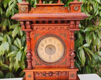 May include: A vintage wooden clock with a detailed, ornate design. The clock features a round face with Roman numerals and decorative metal accents. The clock's wooden frame is a rich, reddish-brown color, and it has a rectangular base.