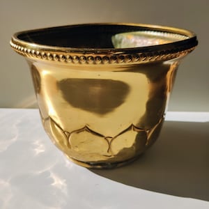 May include: A gold-coloured metal planter with a beaded rim and a decorative pattern of leaves around the bottom.