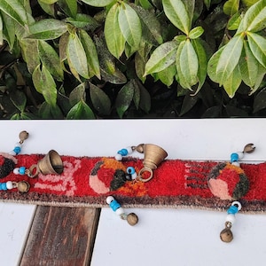 May include: A colourful woven tapestry with bells and beads. The tapestry is red, orange, and blue with a geometric pattern. The bells are brass and the beads are white and blue.