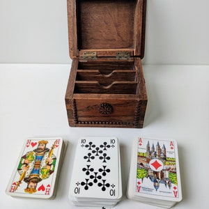 May include: A wooden box with three compartments for storing playing cards. The box is open and three decks of cards are visible. The cards are from a German deck and feature images of cities and buildings. The cards are in the suits of hearts, clubs, and diamonds.