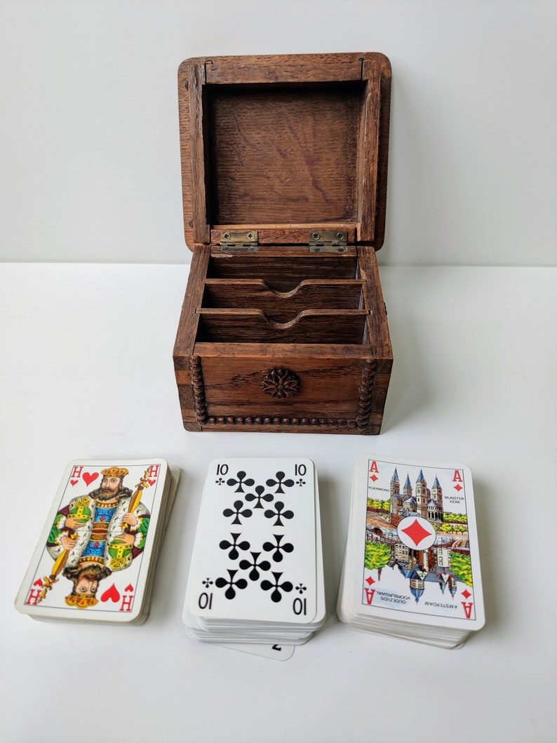 May include: A wooden box with three compartments for storing playing cards. The box is open and three decks of cards are visible. The cards are from a German deck and feature images of cities and buildings. The cards are in the suits of hearts, clubs, and diamonds.
