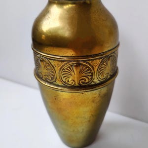 Könnte beinhalten: Eine goldene Metallvase mit einem verzierten Design um die Mitte. Die Vase ist hoch und schmal, mit einem abgerundeten Boden und einem leicht ausgestellten oberen Teil.