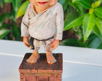 Könnte beinhalten: Eine skurrile Figur eines lächelnden, kahlen Charakters mit rotem Mund und weißem Gewand, der auf einem Holzblock mit dem Buchstaben "J" steht. Die Figur hat einen schwarzen Gürtel und steht vor einem Hintergrund aus grünem Laub.
