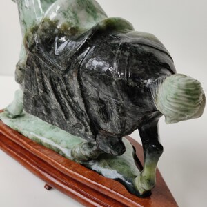 May include: A green and black jade sculpture of a person kneeling on a wooden base. The sculpture is highly detailed and has a glossy finish.